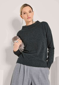 Frau trägt einen dunkelgrünen Pullover und graue, plissierte Hosen, hält eine kleine taupefarbene Clutch und trägt hängende Ohrringe sowie Ringe.