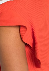 Oranssinauha kangas, jossa on levenevä hiha. Lähikuva, joka näyttää tekstuurin ja muodon, korostaen pehmeää ja kevyttä muotoilua.