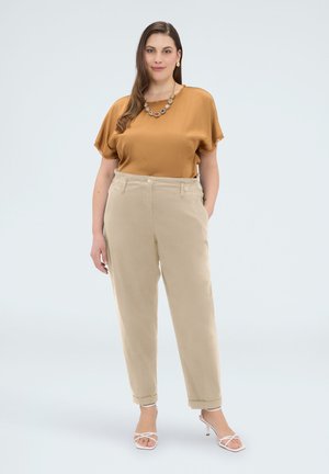 Kvinde stående iført en sennepsfarvet bluse med korte ærmer, beige bukser med høj talje, hvide hæle og en perlehalskæde mod en hvid baggrund.