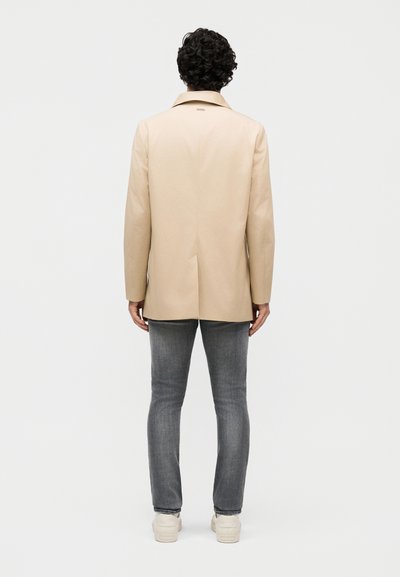 Person med krøllet mørkt hår iført en beige blazer, grå jeans og hvide sneakers, stående med ryggen mod en ensfarvet baggrund.