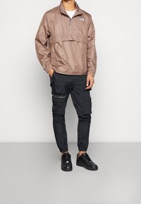 Brązowa, lekka kurtka zapinana na zamek z wysokim kołnierzem, noszona na czarnych spodniach cargo z kieszeniami i zamkami. Czarne sneakersy dopełniają stylizację.