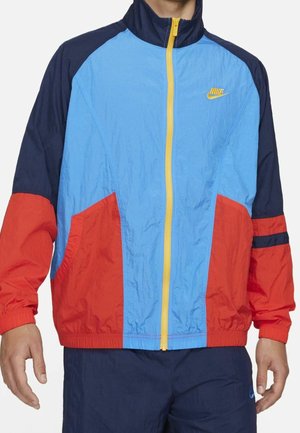 Mężczyzna ubrany w wiatrówkę w kolorowe bloki z panelami w kolorze niebieskim, czerwonym i granatowym, żółtym zamkiem błyskawicznym oraz żółtym logo Nike na piersi.
