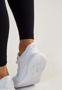 Weißer Sportschuh aus strukturiertem Material, mit runder Zehenpartie und geriffelter Sohle, kombiniert mit schwarzen Leggings, die ein Logo am Saum haben.