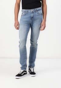 Ljust blå denimjeans med rak passform, lätt slitage på vänster lår och avslutad nederkant. Kombinerat med svarta och vita sneakers.