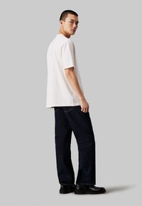 Λευκό κοντομάνικο μπλουζάκι, χαλαρή εφαρμογή, συνδυασμένο με σκούρα μπλε τζιν κατευθείαν γραμμής και μαύρες μπότες στον αστράγαλο, σε φόντο γκρι.