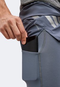 Χέρι τοποθετεί ένα μαύρο smartphone στην γκρι υφασμάτινη πλαϊνή τσέπη αθλητικού σορτς με ετικέτα FORMOTO.