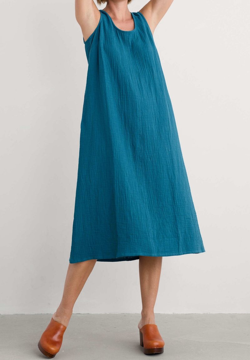 Femme portant une robe midi sans manches en teal avec un tissu texturé et des sabots en bois marron, se tenant les bras levés devant un fond uni.
