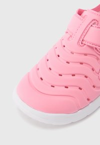 Rožinės gumos avalynė su bangelių tekstūra, perforuotais detalėmis ir balta padais. Yra reguliuojama dirželio sistema, užtikrinanti tvirtą pritvirtinimą.