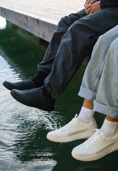 Zapatilla negra con superficie texturizada y suela de goma al lado de una zapatilla blanca de cuero suave, ambas combinadas con pantalones casuales y calcetines.