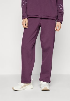 Personne portant un pantalon de survêtement violet et un sweat assorti avec des chaussures de sport blanches, debout devant un fond blanc uni.
