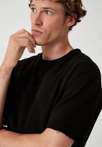 Sort bomulds t-shirt med rund hals og korte ærmer. Har rå kanter ved ærmerne. Modellen poserer eftertænksomt med hånden på hagen.
