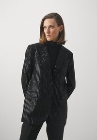Melna sequīnu bleizeris ar vertikālām svītrām, piegrieztiem lakatiem, divām pogām un priekšējām kabatām, uzvilkts virs melna turtlenecka.