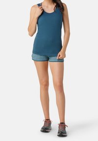 Canottiera blu abbinata a pantaloni corti a quadri blu e verdi. Il modello indossa sneakers grigie con lacci arancioni. Tessuto morbido, stile atletico casual.