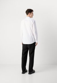 Camisa blanca de manga larga con un dobladillo curvado, combinada con pantalones negros y zapatos negros, vista desde la espalda sobre un fondo liso.