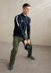 Fekete, hosszú ujjú sportfelső fehér kiegészítőkkel, olívazöld cargo nadrág, szürke sportcipő; egy kerek fekete kettlebellt emel.