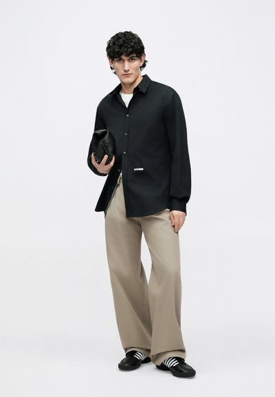 Chemise noire à manches longues avec boutons, pantalon beige à jambes larges, tenant une pochette noire texturée, et portant des chaussures noires avec des bandes blanches.