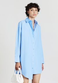 Femme avec des cheveux courts et bouclés portant une robe chemise bleu clair à rayures, tenant un petit sac à main blanc dans sa main gauche.