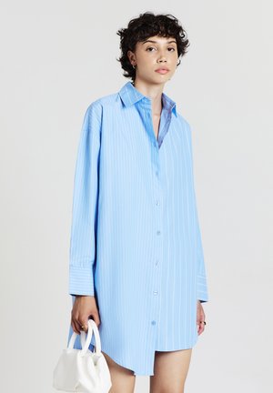 Donna con capelli corti e ricci, che indossa un vestito camicia leggero a strisce blu, tiene una piccola borsa bianca nella mano sinistra.