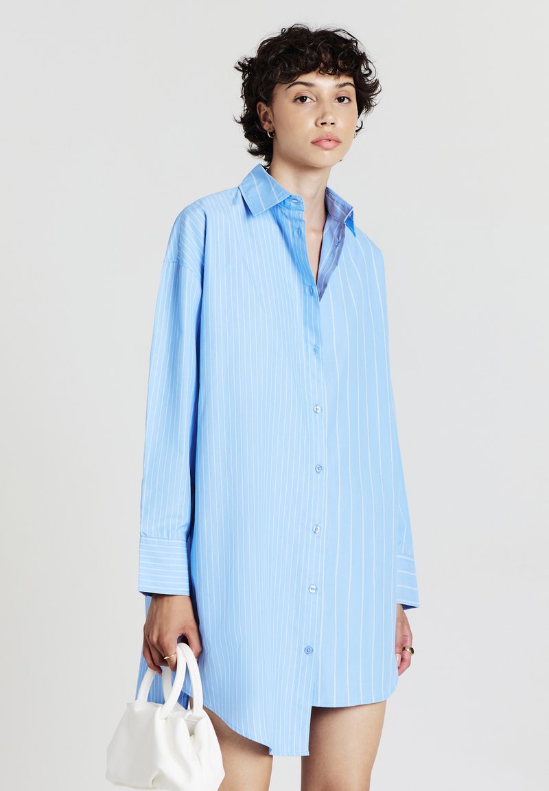 Femme avec des cheveux courts et bouclés portant une robe chemise bleu clair à rayures, tenant un petit sac à main blanc dans sa main gauche.