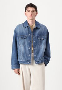 Kék farmerdzseki függőleges fehér csíkkal, két elülső zsebbel, gombos záródással és enyhén oversized kialakítással.