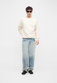 Νέος άνδρας φοράει μαύρα γυαλιά ηλίου, εκρού φούτερ, ανοιχτό μπλε τζιν με μοτίβα λογότυπου και μαύρα παπούτσια, στέκεται με τα χέρια στα πλάγια.