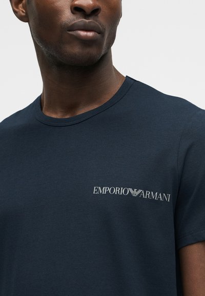 Posnetek od blizu osebe, ki nosi mornarsko modro majico s belim logotipom Emporio Armani na levi strani prsi.