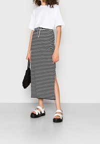 Ženský model v bílém tričku a midi sukni s černobílými pruhy, mašlí v pase a bočním rozparkem, v kombinaci s bílými sandály.