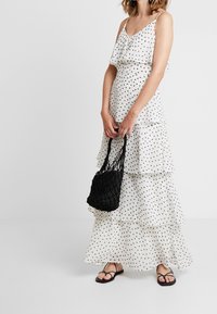 Vestido largo de lunares blancos con volantes en capas, tirantes ajustables, combinado con un bolso negro de mimbre y sandalias negras planas.