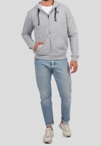 Sudadera gris con cremallera, bolsillo frontal y capucha con cordón, combinada con vaqueros de color azul claro y zapatillas beige. Diseño simple y funcional.