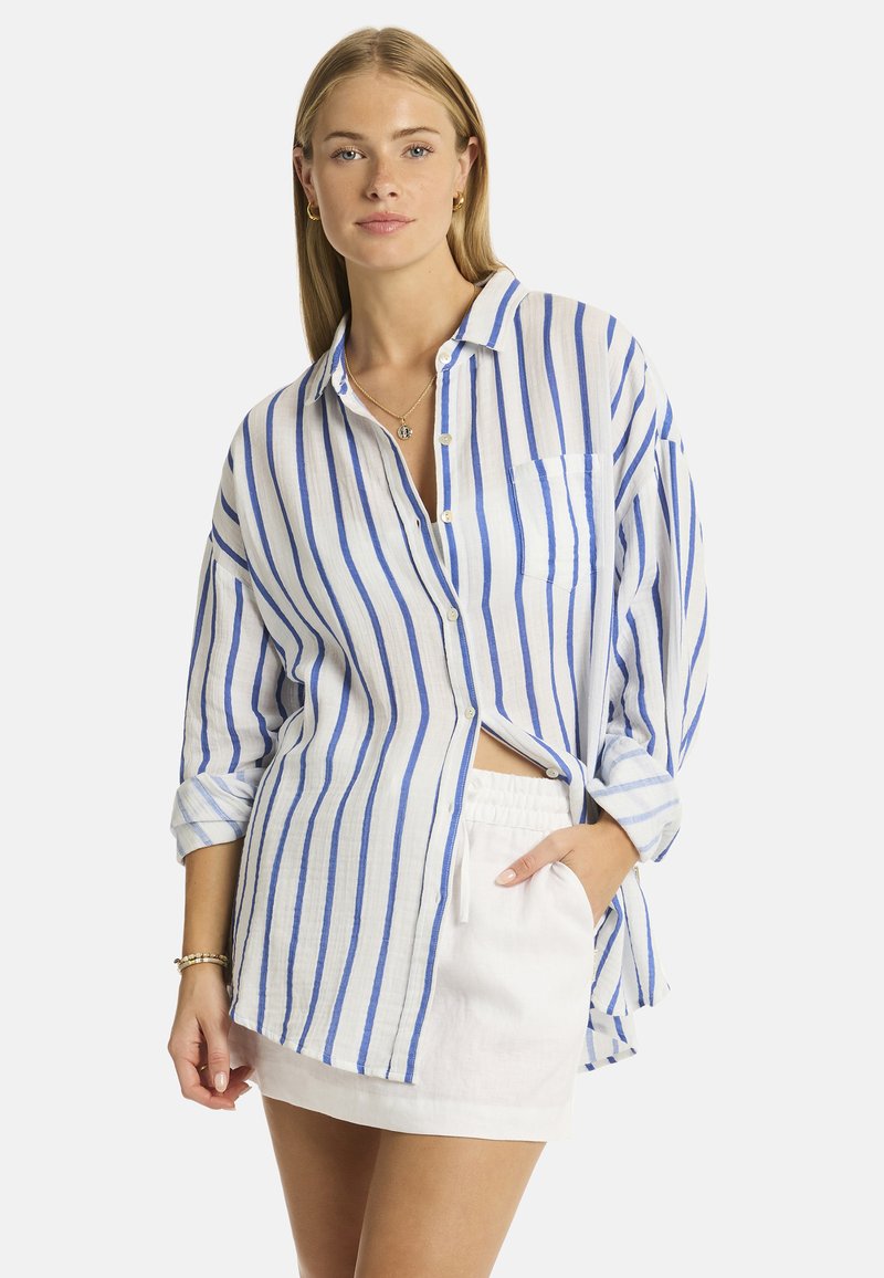 Una donna con lunghi capelli biondi indossa una camicia a righe blu e bianche e pantaloni corti bianchi, posando con una mano in tasca e un'espressione neutra.