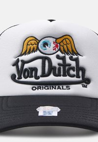 Hvit og svart caps med brodert design som har et øye og vinger, teksten "Von Dutch" i svart, og et lite klistremerke på skyggen.