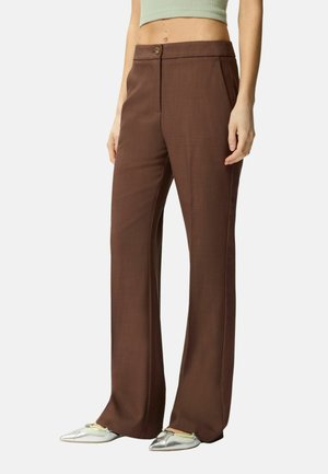Donna che indossa pantaloni marroni a gamba dritta con chiusura a bottoni e ballerine argentate a punta, in piedi davanti a uno sfondo semplice.
