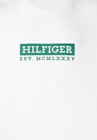 Hvit bomulls T-skjorte med et grønt rektangulært logo som har teksten "HILFIGER" over "EST. MCMLXXXV," med et enkelt, rent design.