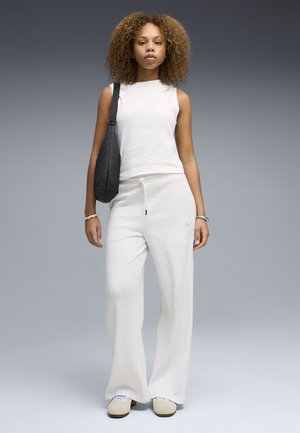 Frau mit lockigem Haar, die ein ärmelloses weißes Oberteil, weiße Hosen mit weitem Bein, beiger Schuhe und eine schwarze Schultertasche trägt, vor grauem Hintergrund stehend.