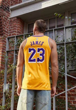 Young man wearing a yellow basketball jersey with "James" and number 23 stands outside near a brick building and metal railing.