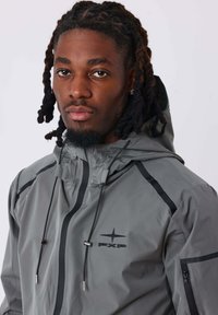 Giacca impermeabile grigia con dettagli neri e cerniere. Presenta cordini regolabili sul cappuccio e un logo sul petto.