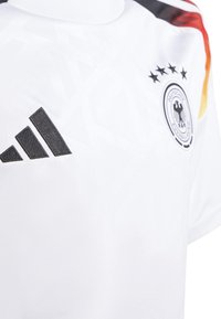 Biała koszulka piłkarska z teksturowanym wzorem. Czarne logo Adidas oraz okrągły emblemat drużyny z podwójnym orłem i gwiazdami.