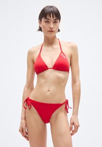 Bikini rosso con top triangolare e slip con laccetti ai fianchi, realizzato in tessuto liscio, con un marchio discreto sulla fascia del top.
