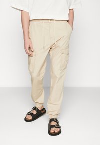 Pantaloni cargo beige con polsini elasticizzati, vita con coulisse e tasche laterali, abbinati a sandali neri con cinghie e fibbie regolabili.