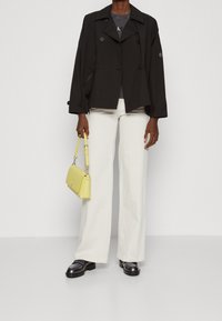 Musta kaksirivinen takki, harmaa t-paita, leveät valkoiset housut, kiiltävät mustat loafers-kengät ja pieni, rakenteellinen muotoiluinen keltainen käsilaukku.