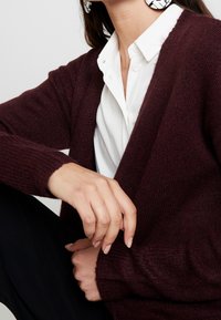 Cárdigan de punto burdeos con puños de canalé, frente abierto y una textura suave, combinado con una camisa blanca de cuello por debajo.