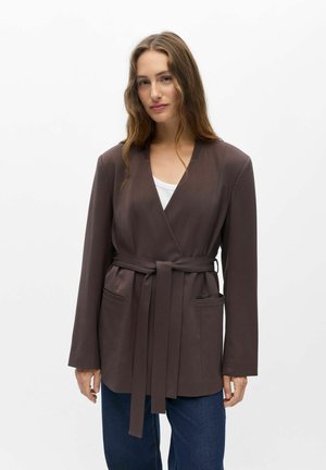 Femme aux longs cheveux bruns portant un blazer marron foncé ceinturé sur un haut blanc et un jean bleu, debout devant un fond blanc.