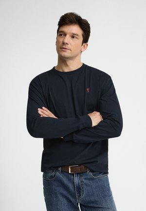 Mann mit dunklen Haaren, trägt ein marineblaues Langarmshirt und Jeans, die Arme verschränkt und schaut leicht zur Seite vor einfachem Hintergrund.