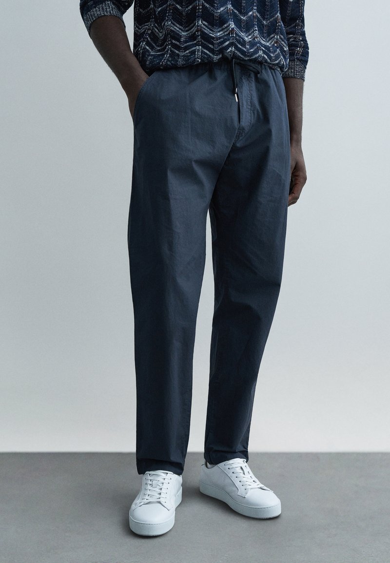 Tamsiai mėlynos lengvos kelnės laisvo kirpimo, su elastinga liemene ir šoninėmis kišenėmis, derinamos su baltais sportbačiais ant pilko paviršiaus.