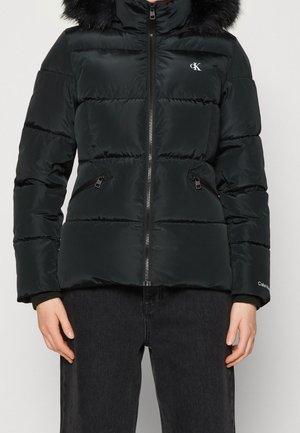 Person wearing a black puffer jacket with a fur-trimmed hood, zipped front, zippered side pockets, and Calvin Klein logos on chest and sleeve.