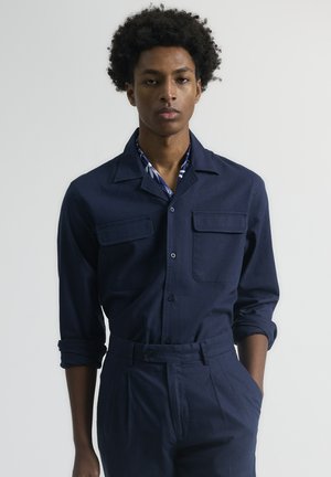 Camisa azul marino de botones, con cuello, dos bolsillos en el pecho y mangas enrolladas, combinada con pantalones a juego, con un ajuste estructurado.
