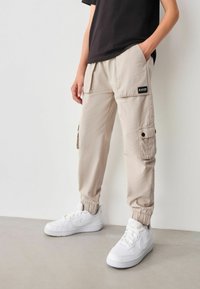 Beige Cargo-Hosen mit elastischen Bündchen, Seitentaschen und einem schwarzen Etikett. Kombiniert mit weißen Sneakers auf einem grauen Boden.