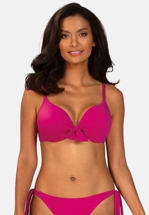 Kobieta o długich, ciemnych włosach, ubrana w jasnoróżowy bikini z kokardkami na górze i wiązanymi bokami na dole, uśmiechająca się do kamery.