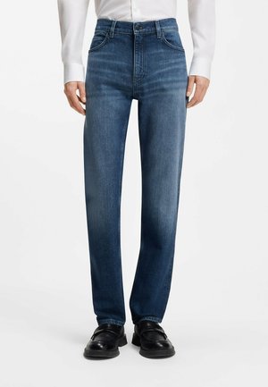 Mężczyzna noszący niebieskie dżinsy o prostych nogawkach z przednimi kieszeniami, połączone z czarnymi skórzanymi butami i częściowo widoczną białą koszulą.