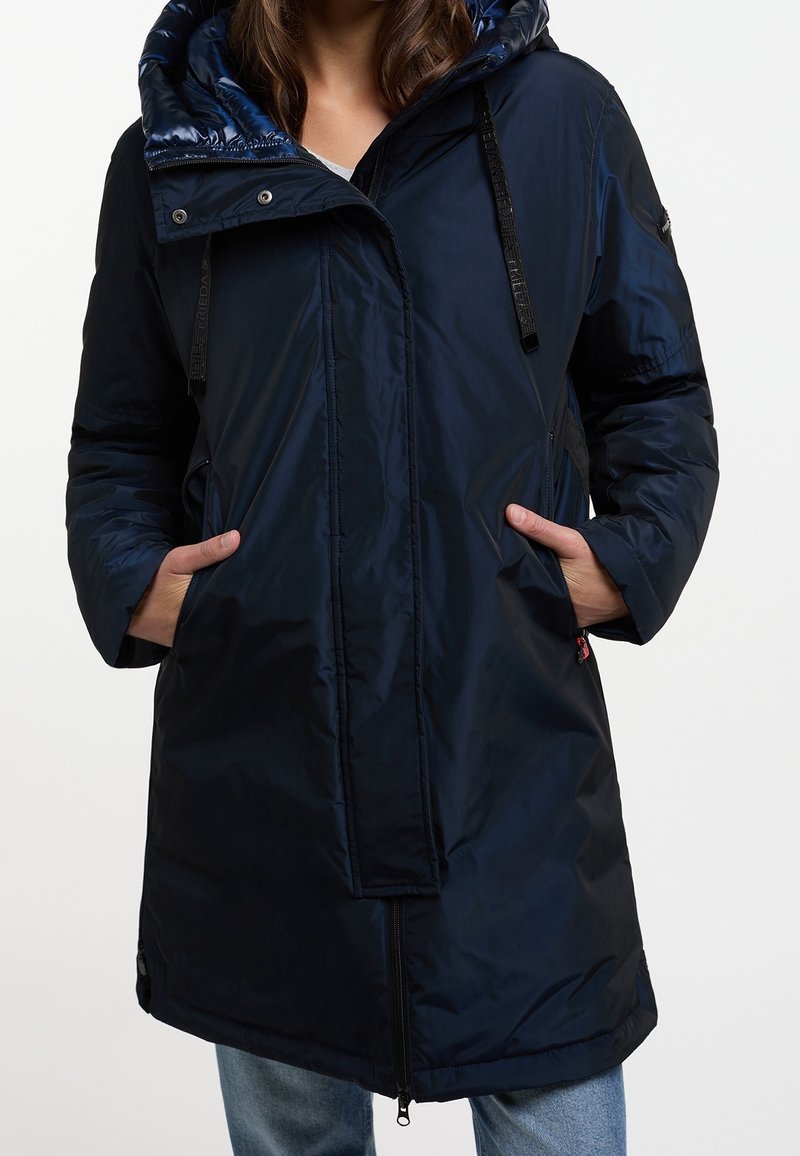 Mujer con una chaqueta larga azul marino acolchada con capucha, manos en los bolsillos y jeans azul claro, parada frente a un fondo blanco liso.